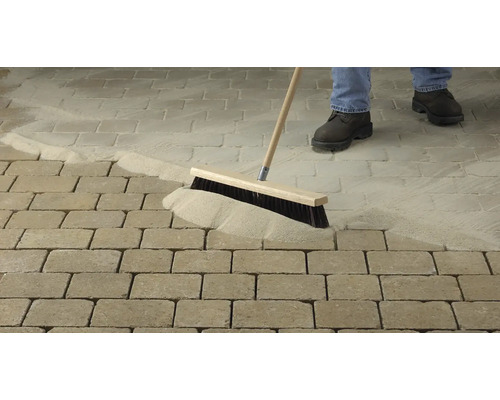 Osoba zametá dlažobný piesok do škár dlaždenej cesty metlou