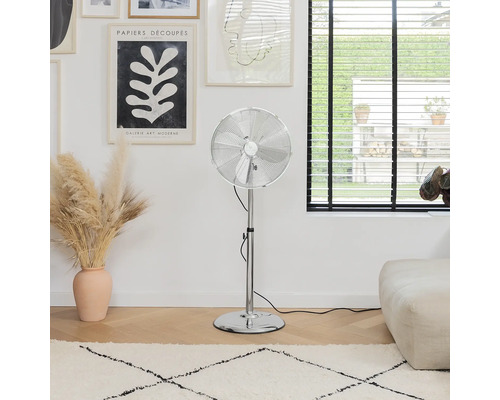 Strieborný ventilátor na podlahe izby s kobercom a umeleckými dielami.