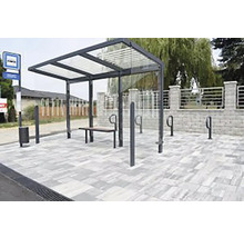 Moderný prístrešok pre cestujúcich s presklenou strechou, kovovým rámom, drevenou lavičkou a stojanmi na bicykle na dláždenej ploche.