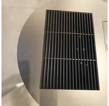Detailný záber na obdĺžnikovú čiernu kovovú ventilačnú mriežku integrovanú do kruhového povrchu z nehrdzavejúcej ocele.