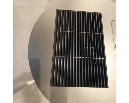 Detailný záber na obdĺžnikovú čiernu kovovú ventilačnú mriežku integrovanú do kruhového povrchu z nehrdzavejúcej ocele.