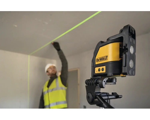 Stavebný robotník v pracovnom odeve označuje stenu pomocou krížového lasera DeWalt na statíve v interiéri.