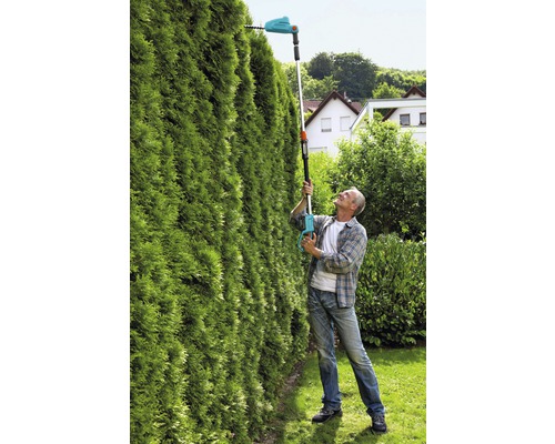 Muž strihá vysoký živý plot akumulátorovými nožnicami na živý plot s teleskopickou tyčou