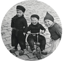 Čiernobiela fotografia troch detí v tradičnom holandskom odeve, jedno jazdí na trojkolke.