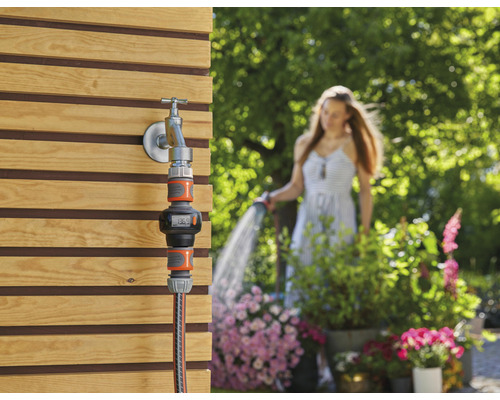 Merač záhradnej vody na vodovodnom kohútiku so ženou v pozadí, ktorá zavlažuje záhradu