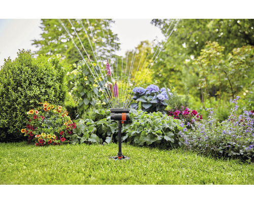 Záhradný postrekovač v prevádzke na trávniku s kvetinovými záhonmi v pozadí.