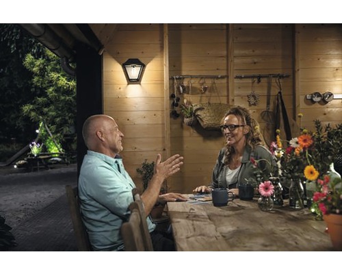 Scéna s mužom a ženou pri drevenom stole pred dreveným domom, osvetlená vonkajšou lampou.