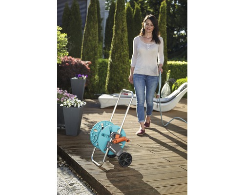 Záhradná scéna so ženou používajúcou záhradnú hadicu na navijaku hadicového vozíka na drevenej terase.