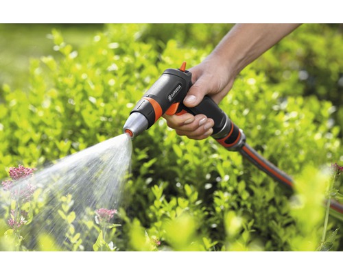 Ruka drží záhradnú striekaciu pištoľ značky Gardena, z ktorej strieka voda na rastliny.