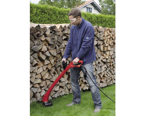 Muž používa vyžínač na trávu v záhrade pred hromadami dreva