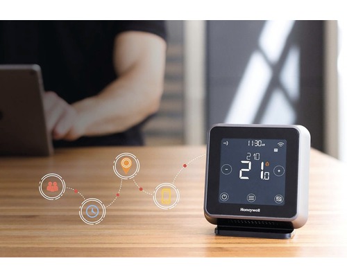 Digitálny termostat Honeywell na stole s osobou v pozadí ovládajúcou tablet