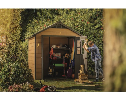 Záhradná scéna so záhradným domčekom, kde je uložené náradie a záhradnícke potreby, a muž rúbe drevo