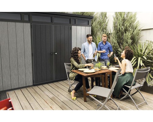 Záhradná scéna s domčekom na náradie a skupinou ľudí, ktorí spolu jedia a pijú.