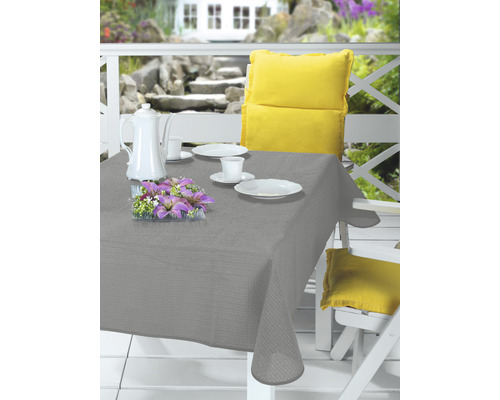 Prestretý záhradný stôl so sivým obrusom, riadom, kávovou kanvicou, kvetinovou dekoráciou a žltými vankúšmi na bielych záhradných stoličkách.