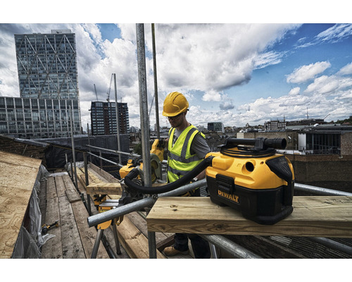 Remeselník s ochrannou prilbou a výstražnou vestou pracuje s mokro-suchým vysávačom Dewalt na lešení pred mestskou scenériou.