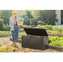 Žena s drevenou debnou a záhradnou lopatkou stojí vedľa otvoreného záhradného boxu v záhrade.