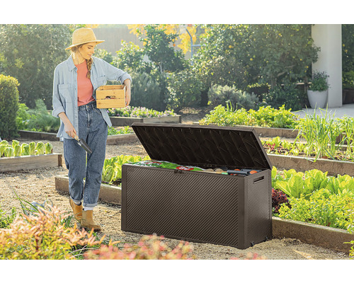 Žena s drevenou debnou a záhradnou lopatkou stojí vedľa otvoreného záhradného boxu v záhrade.