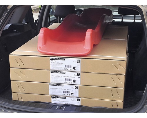 Červený detský šmýkačkový prvok leží na štyroch kartónoch s nápisom Modular Playhouses v kufri auta.