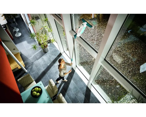 Žena umýva okná čističom okien s teleskopickou tyčou v presvetlenej obývacej izbe.