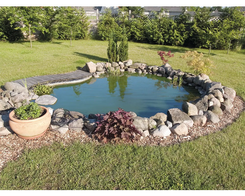 Záhradné jazierko s kamenným obrubníkom a drevenou lávkou v zelenej záhrade.